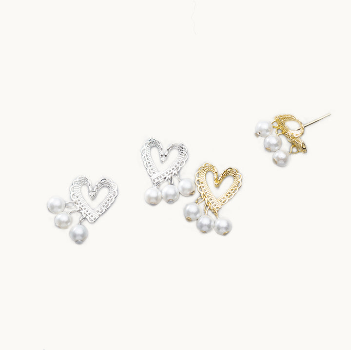 Deux paires de boucles d'oreilles sont exposées devant un fond beige.  Ce sont des boucle d'oreille en forme de coeur. Les coeurs semblent être en dentelle. Des coeurs pendent trois perles. Il y a une paire en argent 925 et une en argent 925 plaqué or.