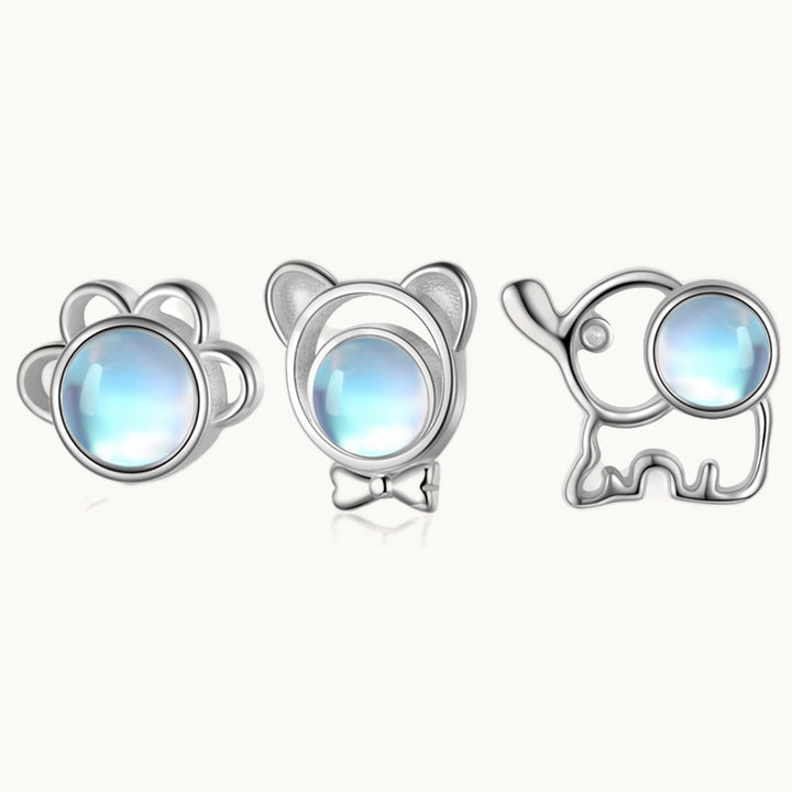 Trois boucles d'oreilles sont exposées devant un fond beige. Il y a une patte de chat, un ourson et un éléphant. Ils ont tous une pierre bleue et sont tous en argent. 