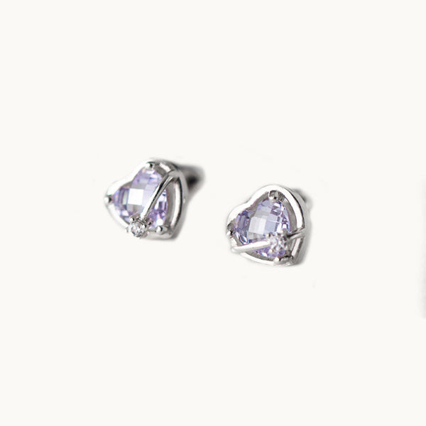 Une paire de boucles d'oreilles est exposée devant un fond beige.  Ce sont des boucles d'oreille en forme de coeur. Elles ont un diamant violet. Il y a une barre fine en métal qui passe devant le coeur, elle est ornée d'un strass. 