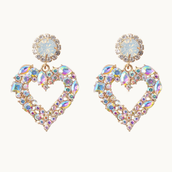 Une paire de boucles d'oreille est exposée devant un fond beige.  Ce sont des boucles d'oreilles imposantes ornées de strass. Elles sont composées d'un diamant rond entouré de petits strass. De ce diamant pend un coeur ajouré entièrement recouvert de strass et de diamants. Ces boucles ont des diamants blancs aux reflets arc-en-ciel .