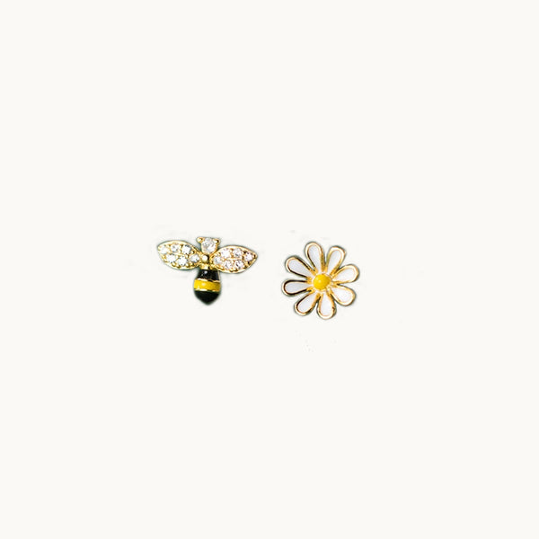 Une paire de boucles d'oreilles est exposée devant un fond beige.  Ce sont des boucles dépareillées : l'une est une fleur et l'autre est une abeille. L'abeille a les ailes et la tête en strass. Elles sont en argent 925 plaqué or.
