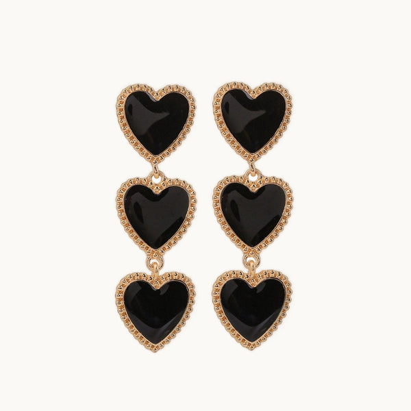 Une paire de boucles d'oreille est exposée devant un fond beige.  Ce sont des boucles d'oreilles pendantes. Elles sont composées de trois coeurs de couleur noire. Les cœurs sont entourés de petites perles métalliques dorées.