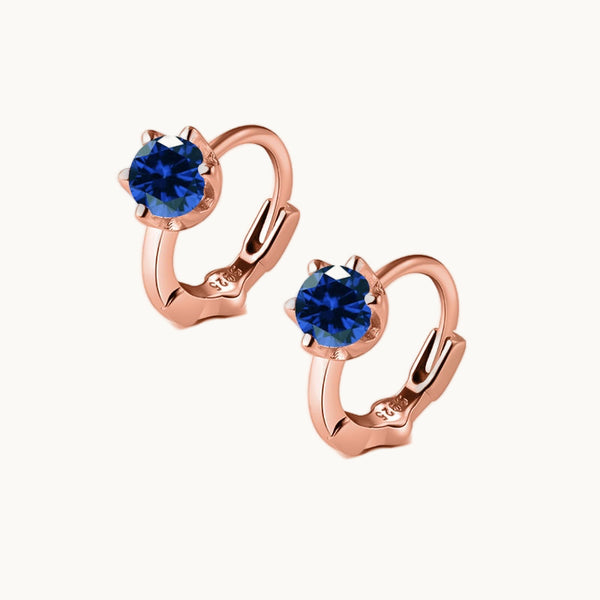 Une paire de boucles d'oreilles est exposée devant un fond beige.  Ce sont des anneaux ornés d'un diamant rond coloré couleur saphir. La boucle est en plaqué or rose. 