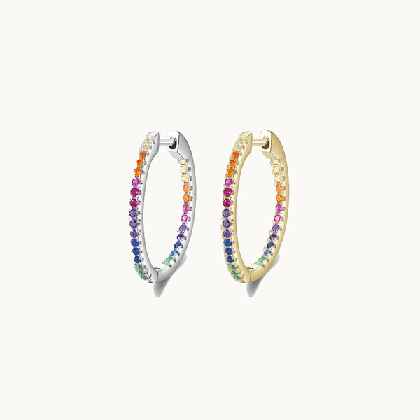 Une paire de boucles d'oreilles est exposée devant un fond beige.  Ce sont des anneaux de style créole ornées de strass colorés. Il y en a une en argent 925 et une en argent 925 plaqué or.