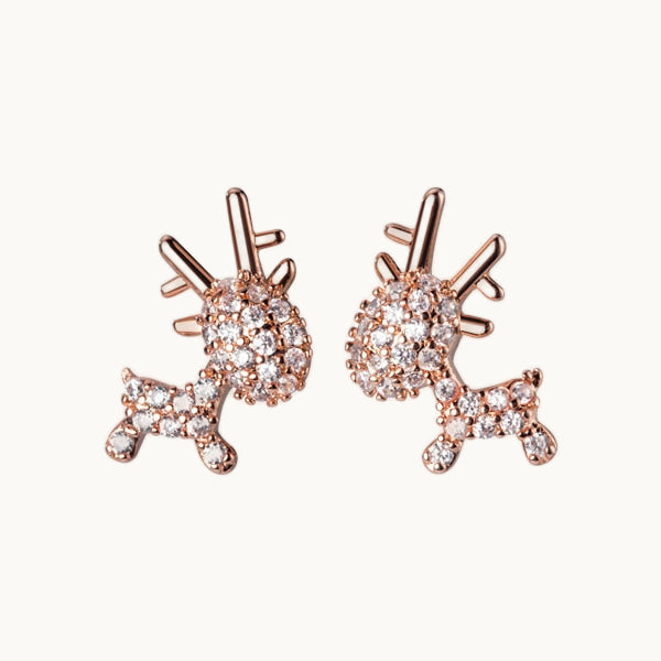 Une paire de boucles d'oreilles est exposée devant un fond beige.  Ce sont des boucles d'oreille en forme de rennes. Les rennes sont entièrement ornés de strass sauf leurs bois. Elles sont en argent 925 plaqué or rose