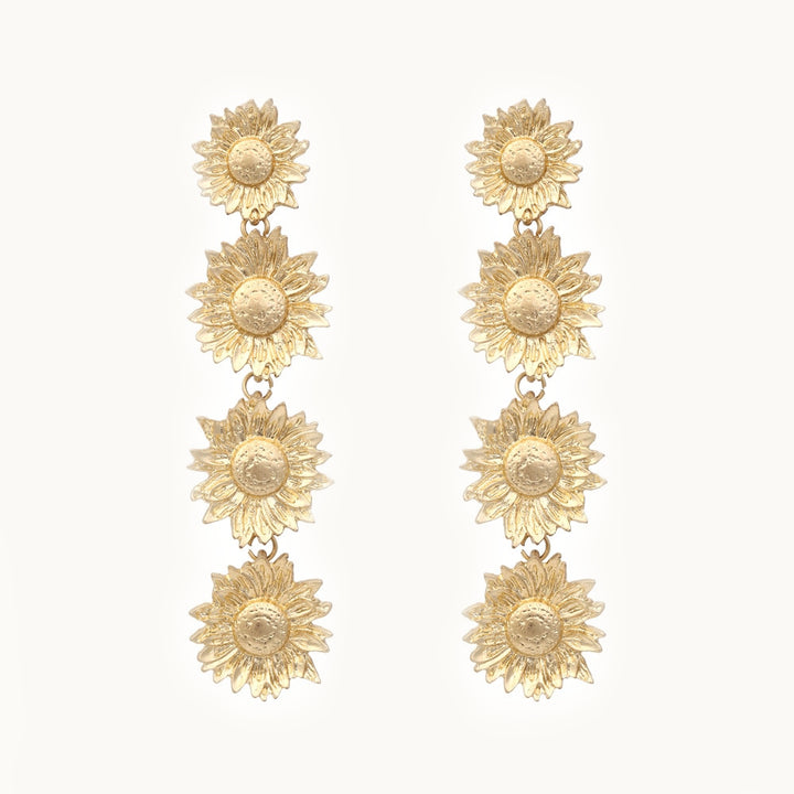 Une paire de boucles d'oreilles est exposée devant un fond beige.  Ce sont des boucles pendantes avec 4 tournesols dorés sur chaque boucle. 