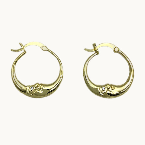 Une paire de boucles d'oreilles est exposée devant un fond beige.  Ce sont des créoles dorées en forme de lune. La lune a un visage et un strass à la place des yeux