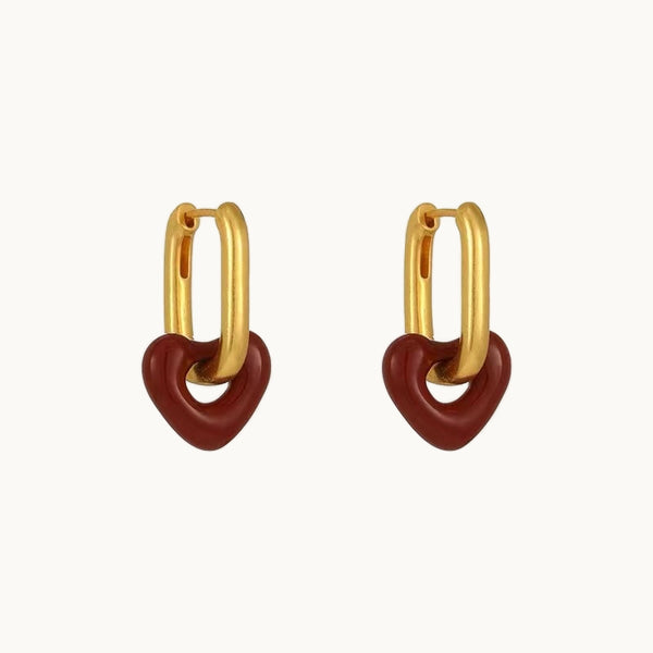 Une paire de boucles d'oreilles est exposée devant un fond beige.  Ce sont des boucles d'oreilles rectangulaires et dorées pendantes sur lesquelles sont suspendues un coeur rouge. 