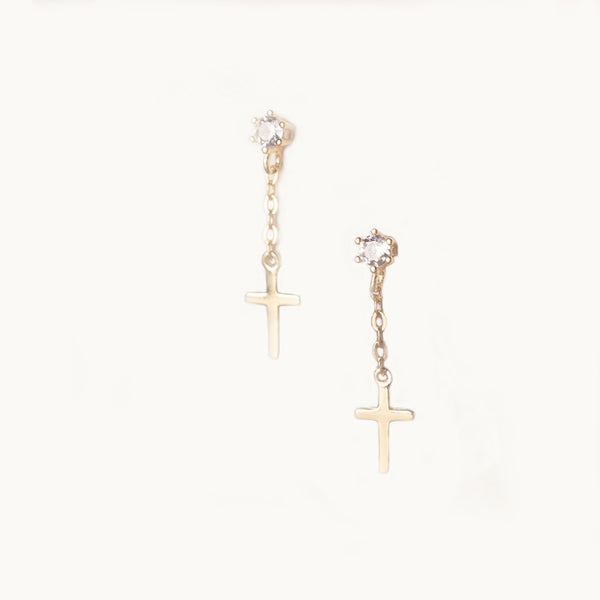 Une paire de boucles d'oreilles est exposée devant un fond beige.  Ce sont des diamants avec une chaîne pendante et une croix au bout. Ce sont des boucles en or.