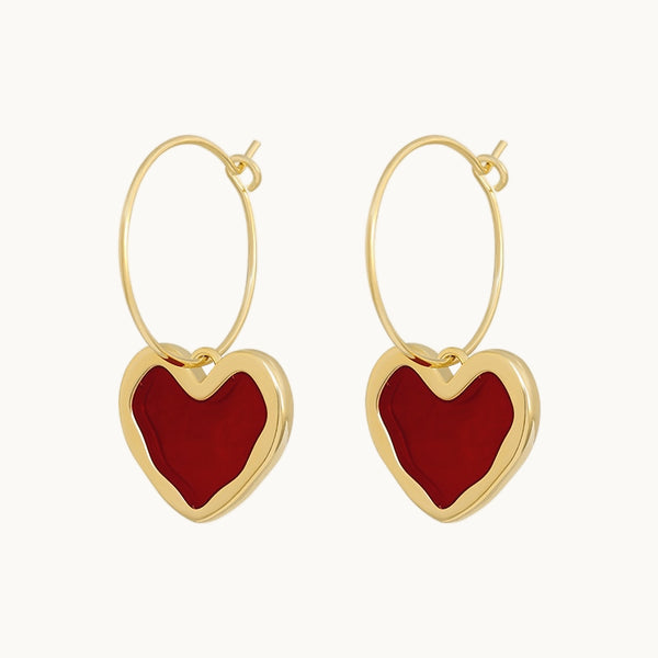 Une paire de boucles d'oreilles est exposée devant un fond beige.  Ce sont des créoles dorées avec un coeur rouge suspendu vintage.