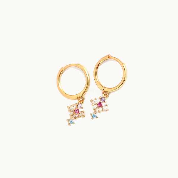 Une paire de boucles d'oreilles est exposée devant un fond beige.   Ce sont des anneaux en or sur lesquelles pend une croix ornée de strass multicolores. 