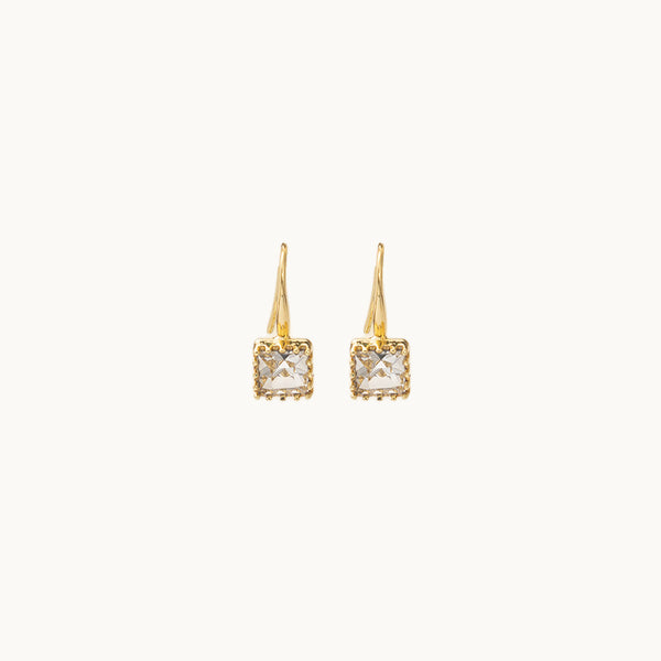Une paire de boucles d'oreilles est exposée devant un fond beige.  Ce sont des boucles d'oreille dormeuses dorées ornées d'un diamant de zirconium carré. 