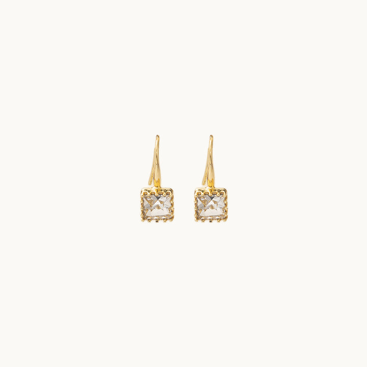 Une paire de boucles d'oreilles est exposée devant un fond beige.  Ce sont des boucles d'oreille dormeuses dorées ornées d'un diamant de zirconium carré. 