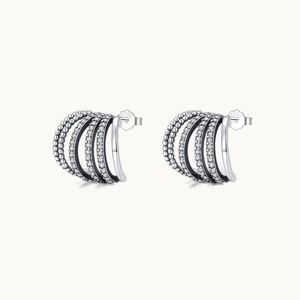 Une paire de boucles d'oreilles est exposée devant un fond beige.  Ce sont des boucles d'oreilles constituées de multiples anneaux non fermées à l'arrière. Les anneaux sont de différentes textures : perles métalliques, anneaux lisses ou encore à strass. Les boucles sont larges. Elles sont en argent 925. 