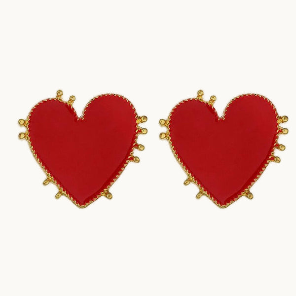 Une paire de boucles d'oreille est exposée devant un fond beige.  Ce sont des boucles d'oreilles en forme coeur de couleur rouge. Le cœur est entouré de petites perles métalliques dorées.