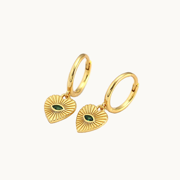 Une paire de boucles d'oreilles est exposée devant un fond beige.  Ce sont des anneaux sur lesquelles pend un coeur vintage avec une émeraude taillée en amande au centre. 
