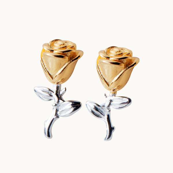 Une paire de boucles d'oreilles est exposée devant un fond beige.  Ce sont des boucles d'oreille en forme de rose avec la tige. La tige est en argent 925. La rose est en argent 925 et en argent 925 plaqué or. 