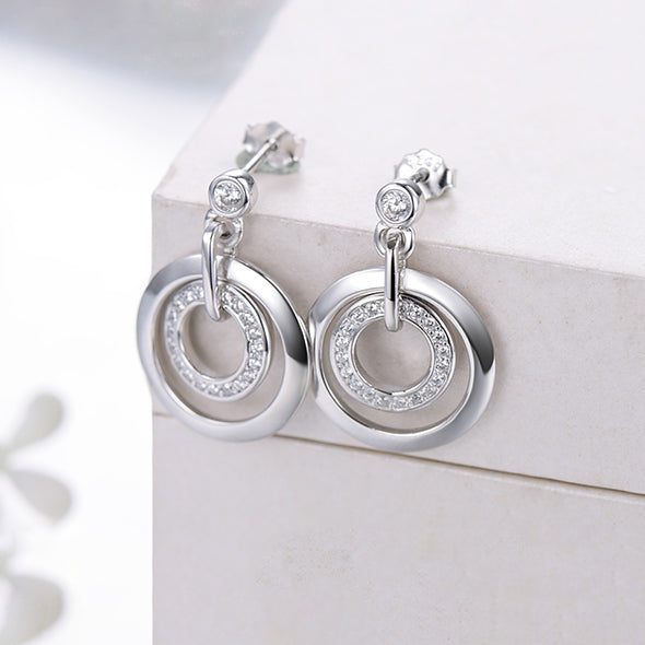 Une paire de boucles d'oreille pendantes en argent 925 avec des diamants, deux anneaux interconnectés, et un petit anneau incrusté de strass. Élégance et raffinement pour chaque occasion.