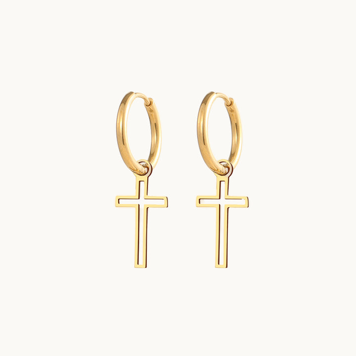 Une paire de boucles d'oreilles est exposée devant un fond beige. Ce sont des boucles d'oreille constituées d'un anneau fin sur lequel pend une croix ajourée. Elles sont en acier inoxydable plaqué or.  