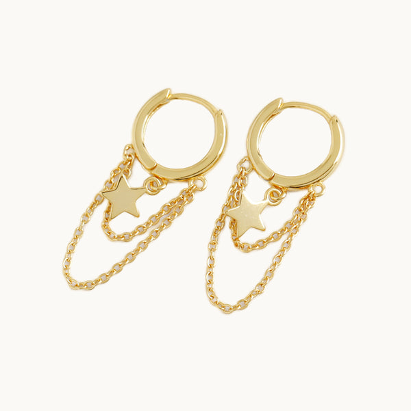 Une paire de boucles d'oreilles est exposée devant un fond beige.  Ce sont des anneaux sur lesquelles pendent des chaînes et des étoiles.