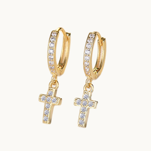 Une paire de boucles d'oreilles est exposée devant un fond beige.  Ce sont des boucles d'oreille anneau avec une croix pendante. Elles sont ornées de strass sur l'anneau et sur la croix. Elles sont en argent 925 plaqué or.