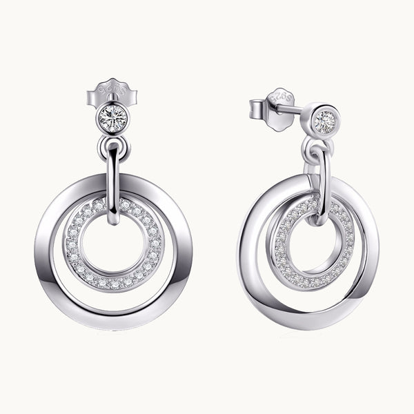 Une paire de boucles d'oreilles est exposée devant un fond beige.  Ce sont des boucles d'oreille composées d'un petit diamant rond sur lequel sont suspendus deux anneaux : le plus petit est orné de strass et est entouré par un plus gros anneau qui est en argent 925.