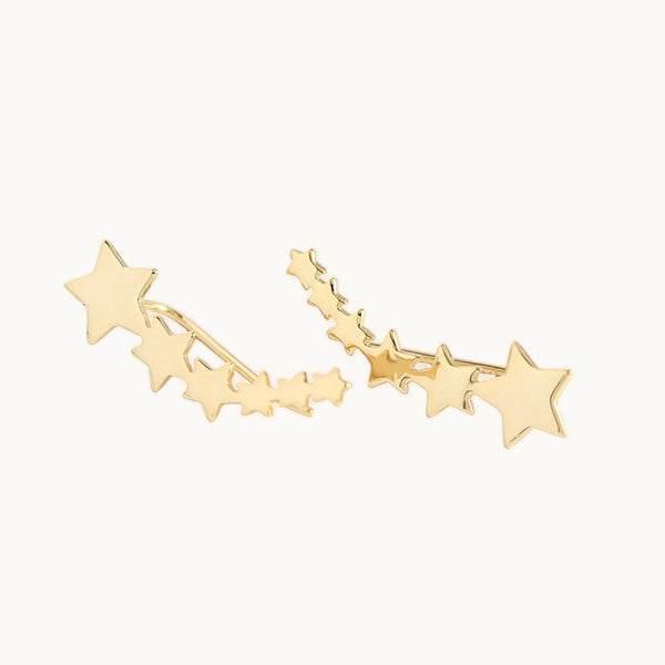 Une paire de boucles d'oreilles est exposée devant un fond beige.  Ce sont des boucles grimpantes avec des étoiles disposées de la plus petite à la plus grande. Elles sont en plaqué or. 