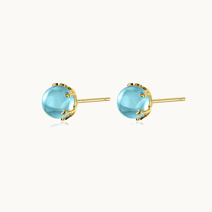 Une paire de boucles d'oreilles est exposée devant un fond beige.  Ce sont des pierres en pierre transparente bleue. La base est en argent 925 plaqué or.