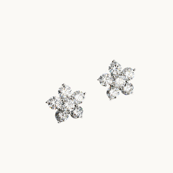 Une paire de boucles d'oreilles est exposée devant un fond beige. Ce sont des boucles d'oreille en forme de fleur. Chaque pétale et le coeur de la fleur sont en diamant moissanite. 