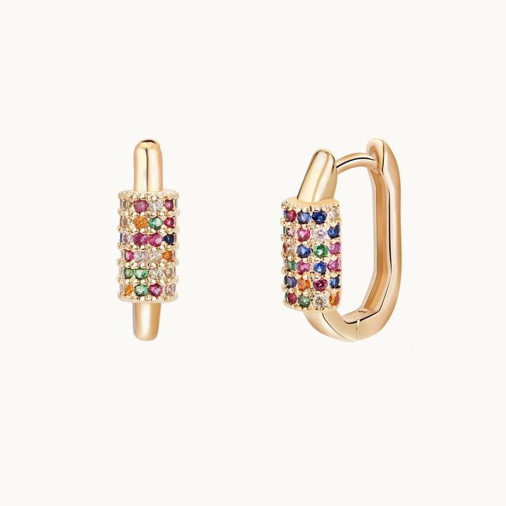 Une paire de boucles d'oreilles est exposée devant un fond beige. Ce sont des créoles ovales dorés ornées d'une bague sur le devant qui est sertie de strass multicolores.