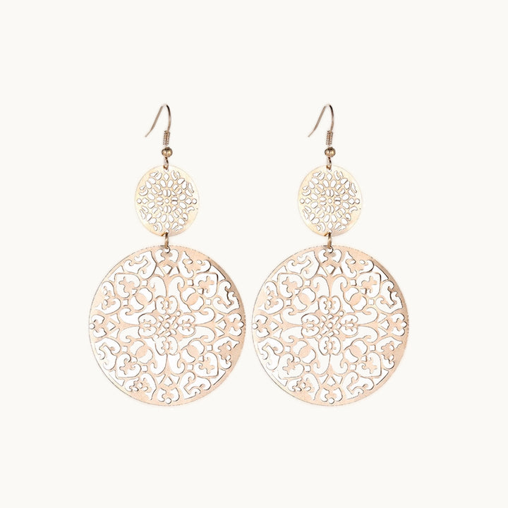 Une paire de boucles d'oreilles est exposée devant un fond beige. Ce sont des boucles d'oreille pendante. Elles sont composées de deux ronds superposés, le premier est plus petit que le second. Les ronds sont comme des mandalas ajourés. Elles sont dorées.