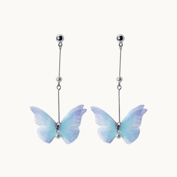 Une paire de boucles d'oreilles est exposée devant un fond beige.  Ce sont des boucles longues et pendantes. Elles ont des perles à plusieurs endroits sur la longueur et il a un papillon bleu au bout d'aile violette.Les papillons sont légers et donne une touche féérique. Elles sont en argent 925. 
