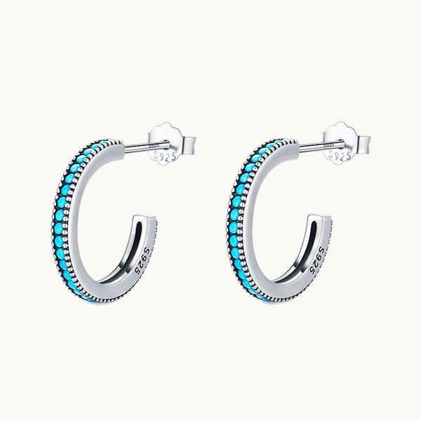 Une paire de boucles d'oreilles est exposée devant un fond beige.  Ce sont des créole de style bohème ornées de petites pierres turquoises tout du long.  Elles sont en argent 925.