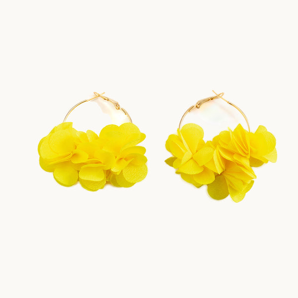 Une paire de boucles d'oreilles est exposée devant un fond beige.  Ce sont des boucles d'oreille créoles dorées fine. Elles sont ornées de fleurs en tissu. Les fleurs sont jaunes.