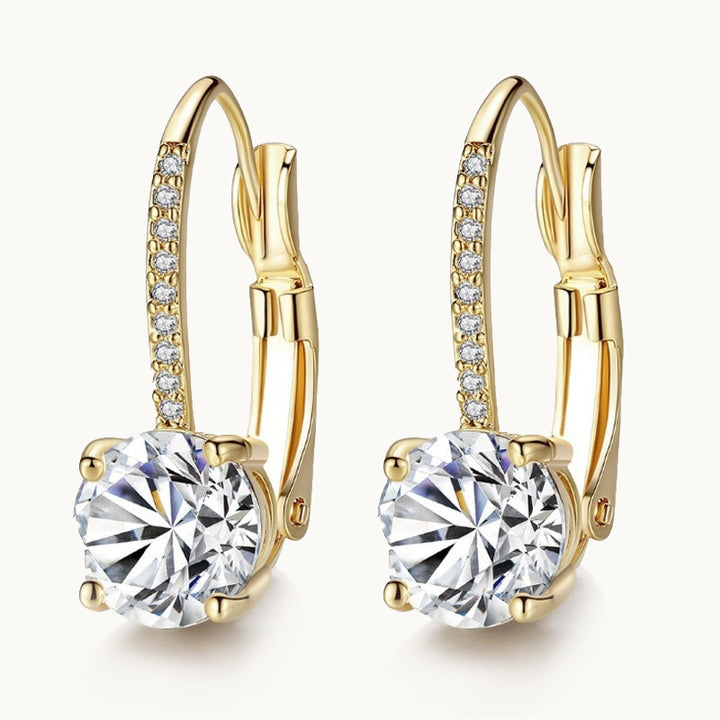 Une paire de boucles d'oreilles est exposée devant un fond beige.  Ce sont des boucles d'oreille dormeuse. Il y a un gros diamant au bout et le devant est orné de petits diamants scintillants en forme de petits strass. Elles sont en argent 925 plaqué or.