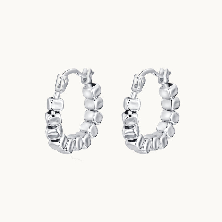 Une paire de boucles d'oreilles est exposée devant un fond beige.  Ce sont des créoles ornées de cubes en argent 925.