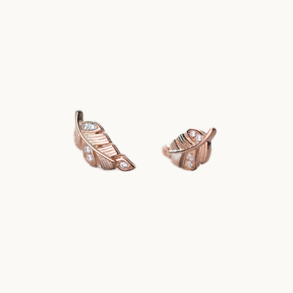 Une paire de boucles d'oreilles est exposée devant un fond beige.  Ce sont des petites plumes ornées de strass. Elles sont en argent 925 plaqué or rose. 
