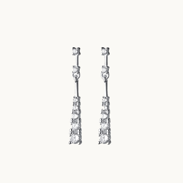 Une paire de boucles d'oreilles est exposée devant un fond beige.  Ce sont des boucles d'oreille pendante ornées de diamants. Elles sont en argent 925.