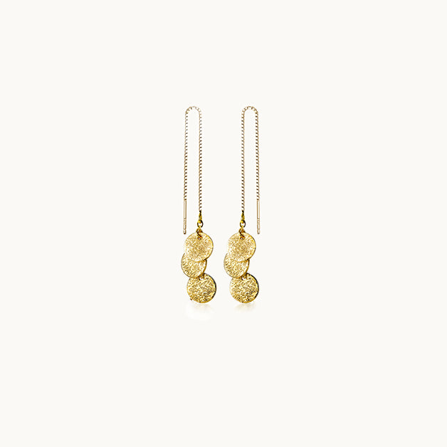 Une paire de boucles d'oreilles est exposée devant un fond beige.  Ce sont des boucles longues. Elles sont constituées d'une chaîne pendante ornée au bout de trois ronds martelés. Elles sont en argent 925 plaqué or.