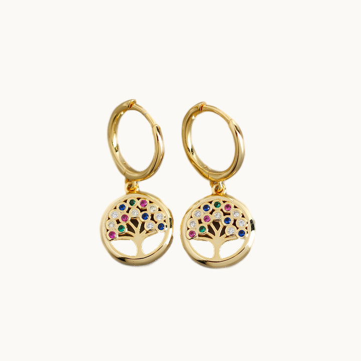 Une paire de boucles d'oreilles est exposée devant un fond beige.  Ce sont des anneaux sur lesquels pend un rond dans lequel il y a un arbre de vie avec strass colorés. 