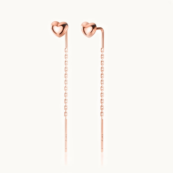 Une paire de boucles d'oreilles est exposée devant un fond beige.  Ce sont des petits coeurs sur lesquels est pendue une chaîne.  Elles sont en or rose.