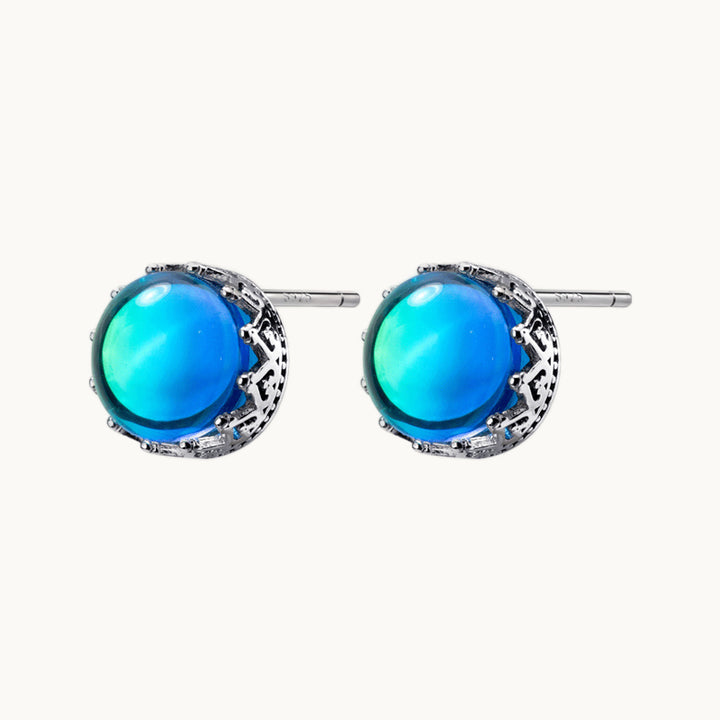 Une paire de boucles d'oreilles est exposée devant un fond beige.  Ce sont des boucles d'oreilles rondes. Elles sont composées d'un cristal rond bleu avec des reflets arc-en-ciel. La pierre est sertie par de l'argent 925 qui a une forme de couronne royale. 