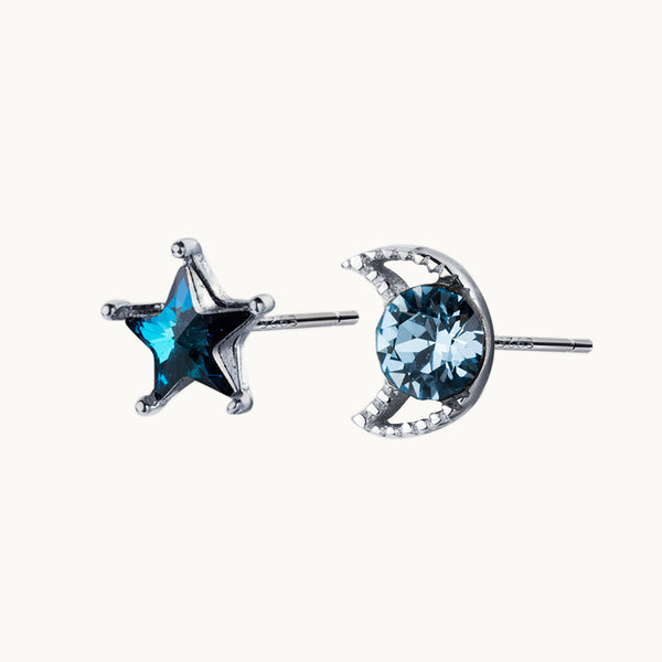 Une paire de boucles d'oreilles est exposée devant un fond beige.  Ce sont des boucles asymétriques : l'une est une étoile et l'autre une lune. Elles sont ornées d'un diamant en cristal bleu saphir. Elles sont en argent 925.