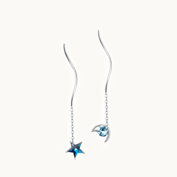 Une paire de boucles d'oreilles est exposée devant un fond beige.  Ce sont des boucles d'oreille pendante. Elles sont constituées d'une chaîne et d'une tige ondulée. Elles sont dépareillées, l'une a une étoile au bout et l'autre une lune. La lune et l'étoile sont ornées d'un diamant de zirconium de couleur bleu saphir. Elles sont en argent 925.
