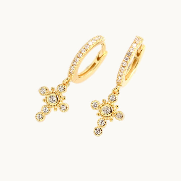 Une paire de boucles d'oreilles est exposée devant un fond beige.  Ce sont des anneaux en strass avec une croix pendante en strass. Ce sont des boucles d'oreille en plaqué or. 