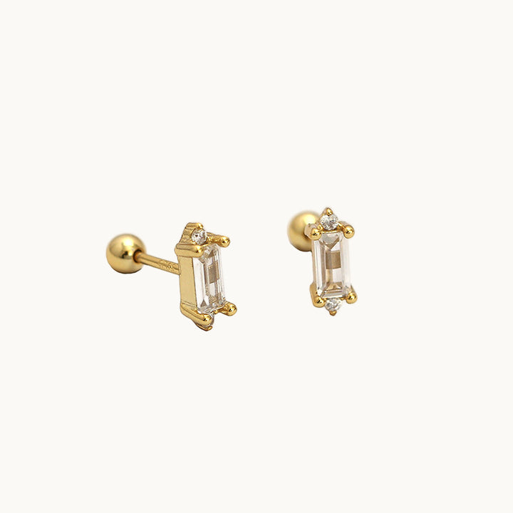 Une paire de boucles d'oreilles est exposée devant un fond beige.  Les boucles sont rectangulaires avec des strass. La tige est une barre droite. Les boucles sont en plaqué or. 