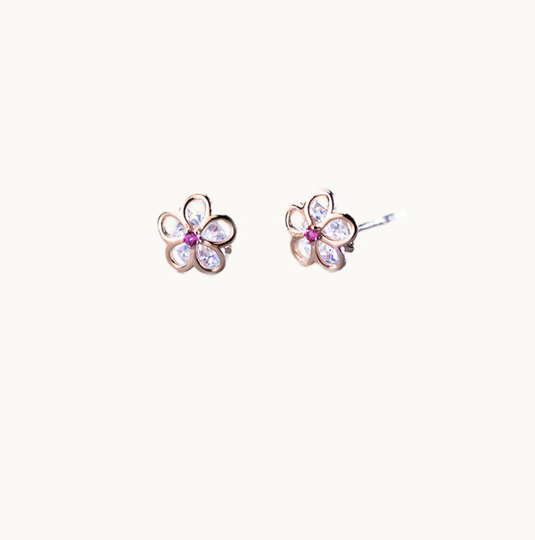 Une paire de boucles d'oreilles est exposée devant un fond beige.  Ce sont des fleurs ajourées en argent 925 or rose. Le milieu de la fleur est un strass rose fuchsia. Au coeur de la fleur ajourée se trouve un gros diamant de zirconium très brillant. 