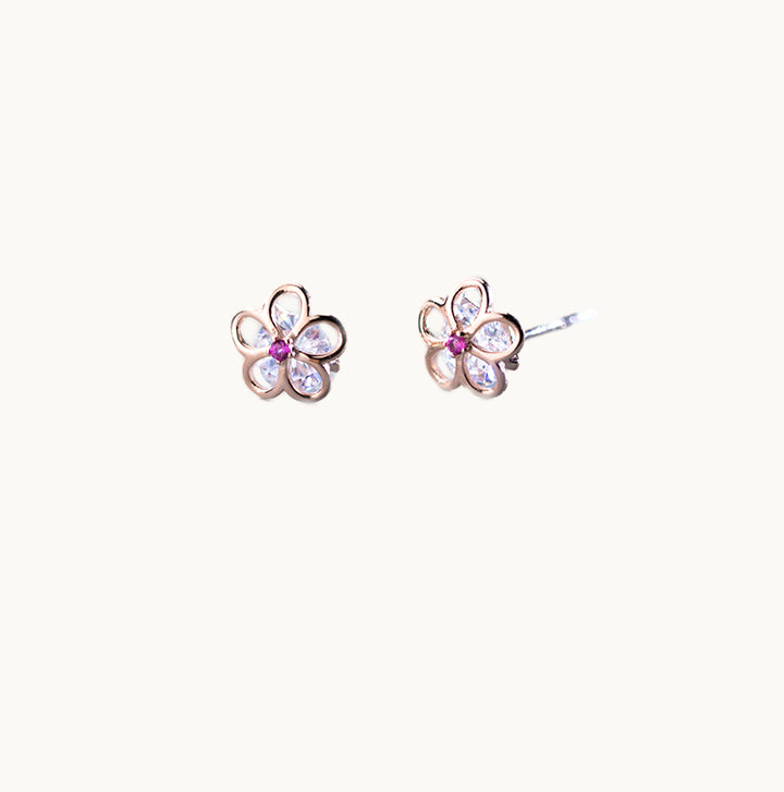 Une paire de boucles d'oreilles est exposée devant un fond beige.  Ce sont des fleurs ajourées en argent 925 or rose. Le milieu de la fleur est un strass rose fuchsia. Au coeur de la fleur ajourée se trouve un gros diamant de zirconium très brillant. 