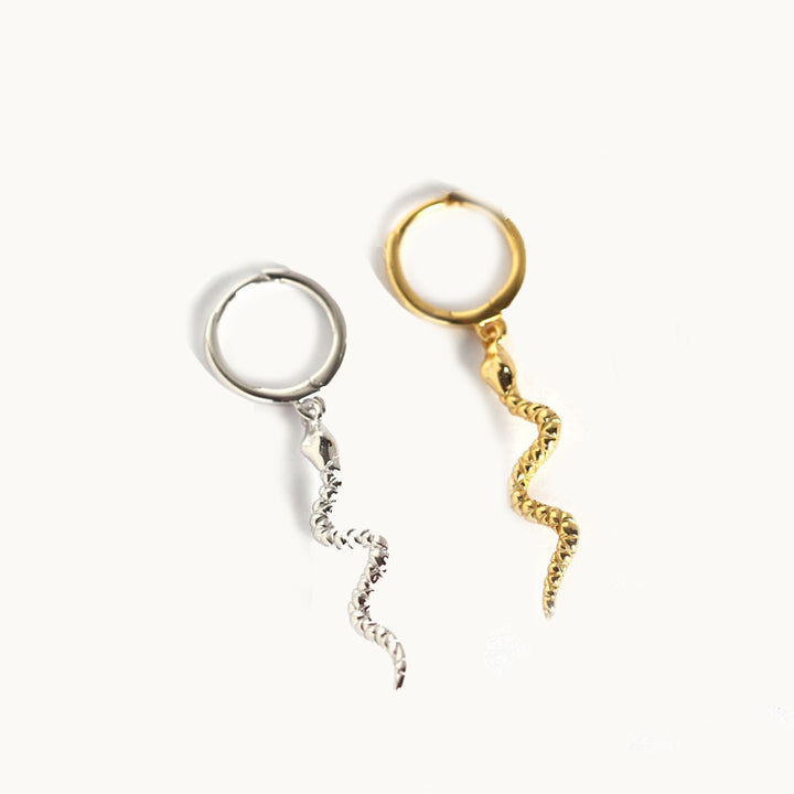 Deux boucles d'oreilles sont exposées devant un fond beige. Ce sont des anneaux sur lesquels pend un serpent. L'une est en or et l'autre en argent. 