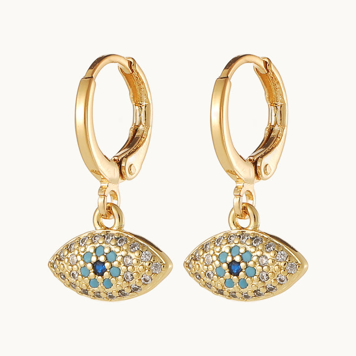 Une paire de boucles d'oreilles est exposée devant un fond beige. Ce sont des boucles d'oreille en forme d'anneau sur lequel pend un oeil doré. L'iris et la pupille sont ornés de strass bleu et noir. Le reste de l'oeil est orné de strass transparents. Elles sont dorées.